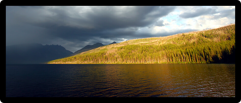 Sunlight illuminates the shoreline of Kintla Lake against a stormy sky in Glacier National Park.
